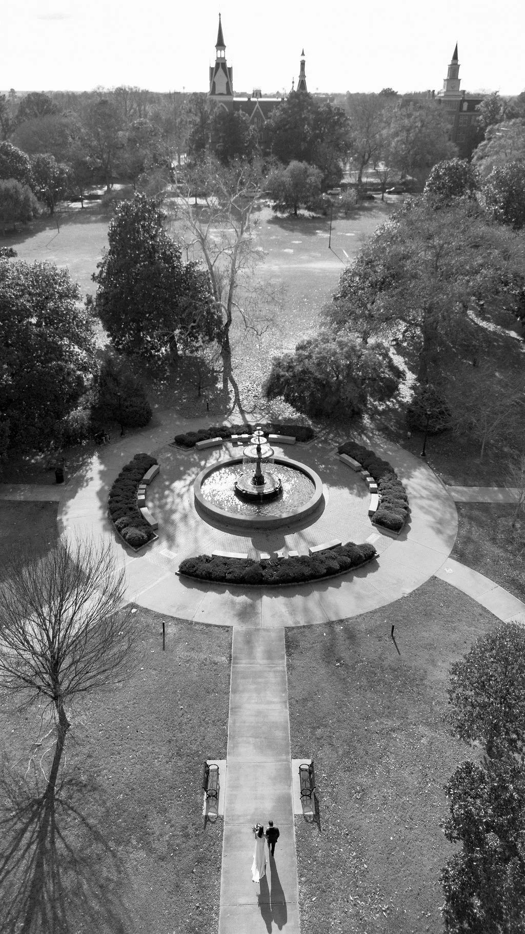 A black and white photo from a high angle showing a park with a fountain at the center, trees, benches, and a wedding couple walking down a path.