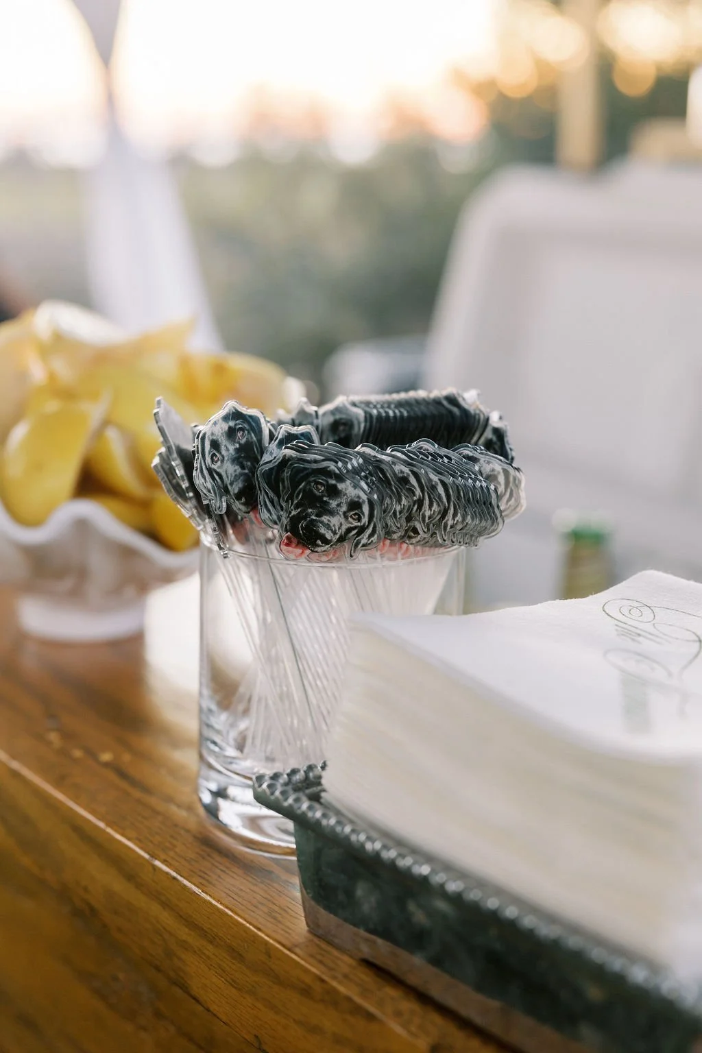 Candy or chocolate in the shape of black Labrador retriever faces in a glass bowl on a wooden table, with a bowl of potato chips in the background, in an outdoor setting during sunset.