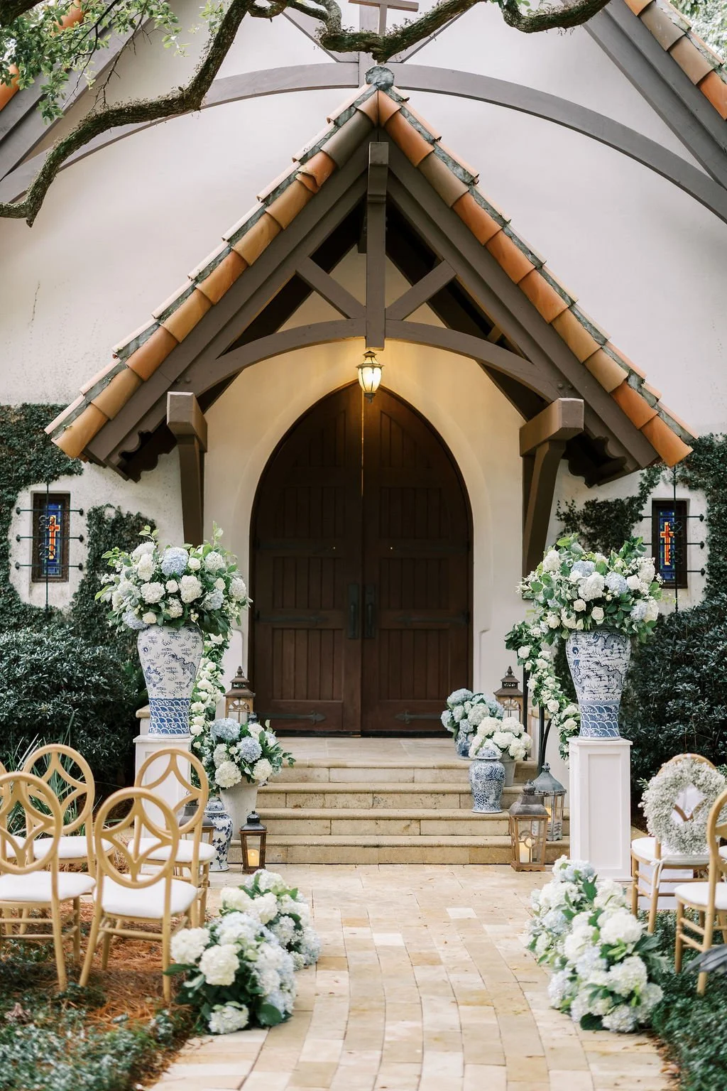 Wedding ceremony setup outside a building with a wooden door, decorated with large floral arrangements of white flowers and greenery, lanterns, and white chairs arranged for guests.