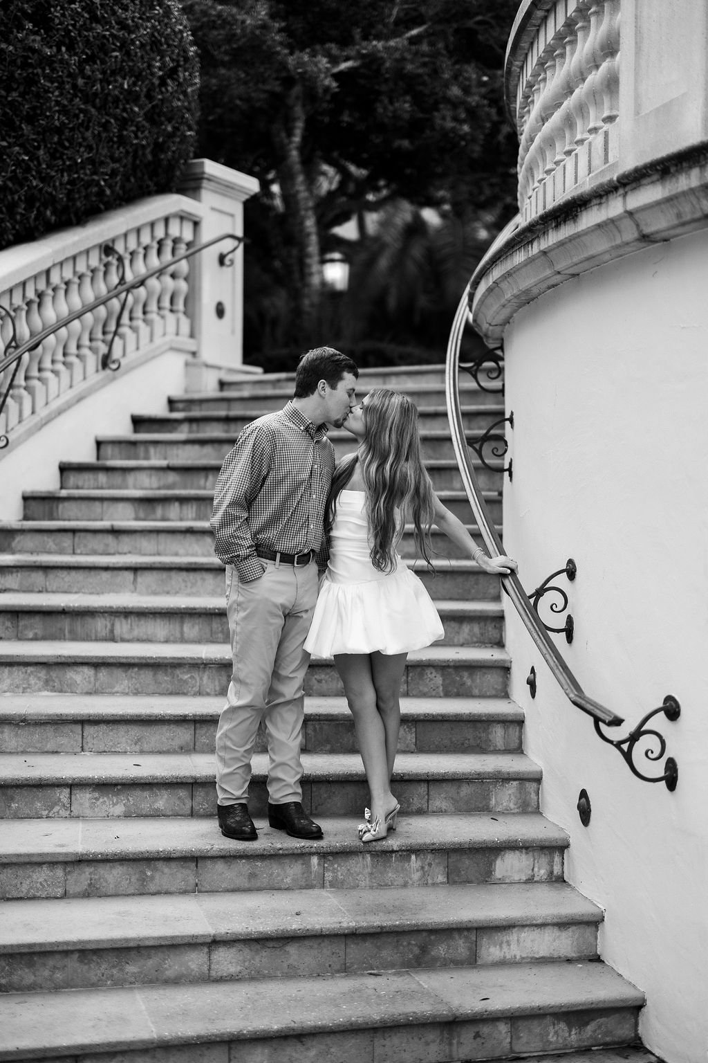 A black and white photo of a young couple kissing on a staircase outside, with the man in a checkered shirt and light-colored pants, and the woman in a short, strapless dress with long hair, holding onto the handrail.
