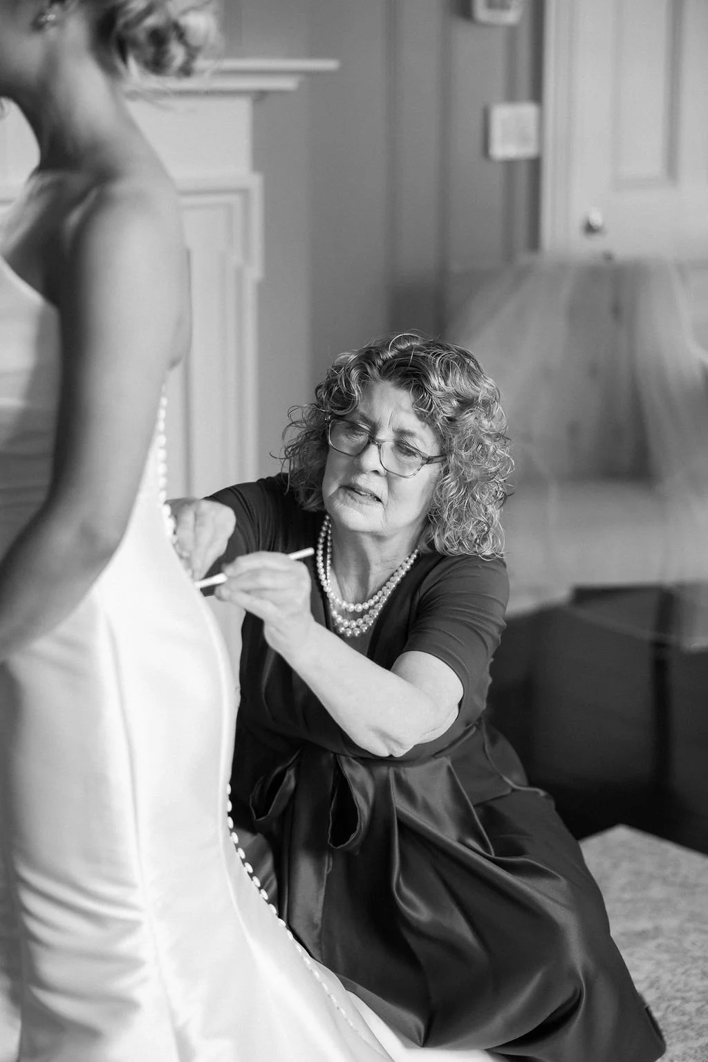 A woman with curly hair and glasses, dressed in a dark top with pearl necklace, is meticulously sewing beads onto a bridal gown, which is on a mannequin, in a room with wooden flooring and a closed door in the background.