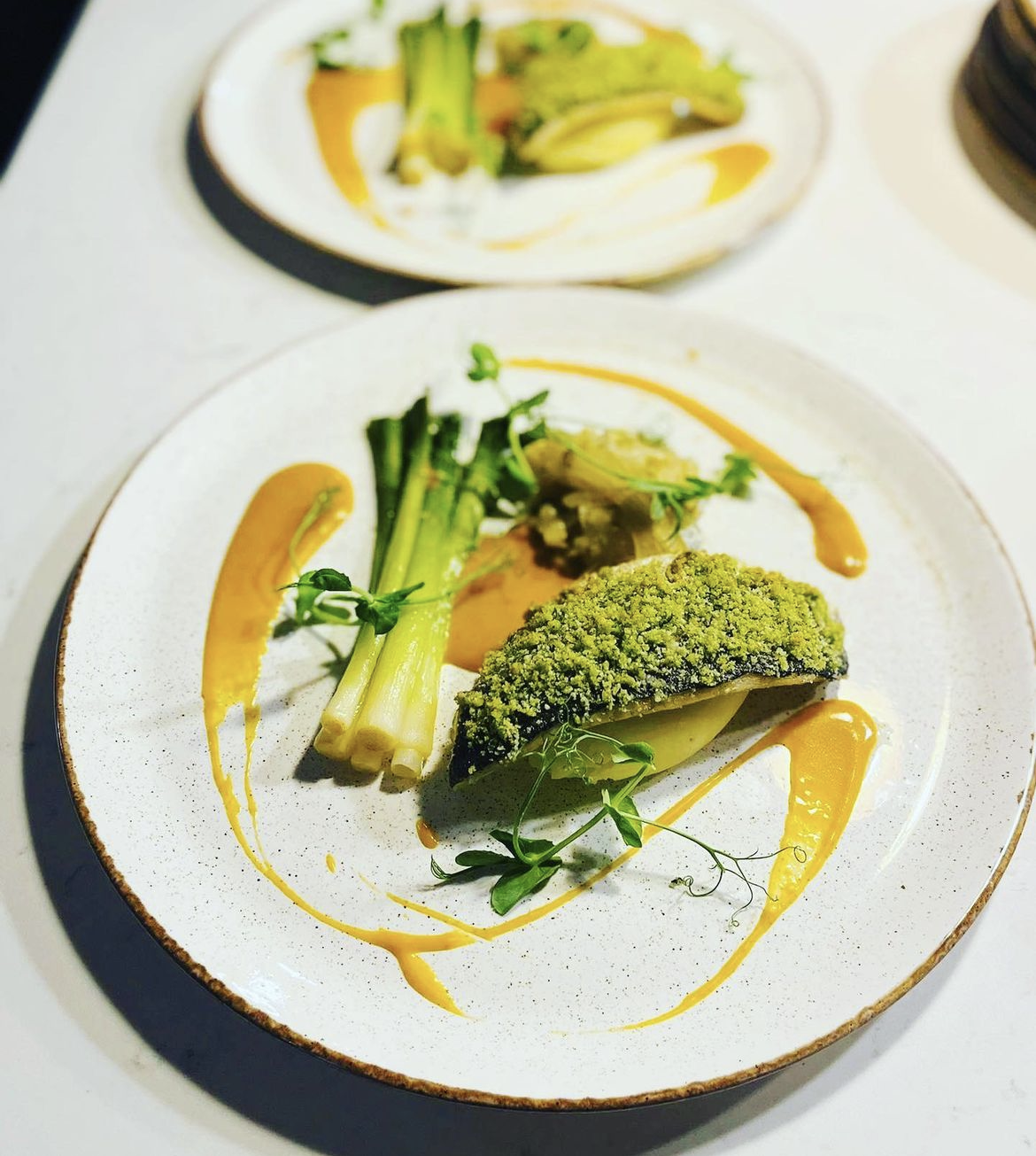 A plated fish dish featuring catch of the day with a tarragon crust, seasonal vegetable garnish and lobster bisque sauce artfully drizzled around on a white plate.