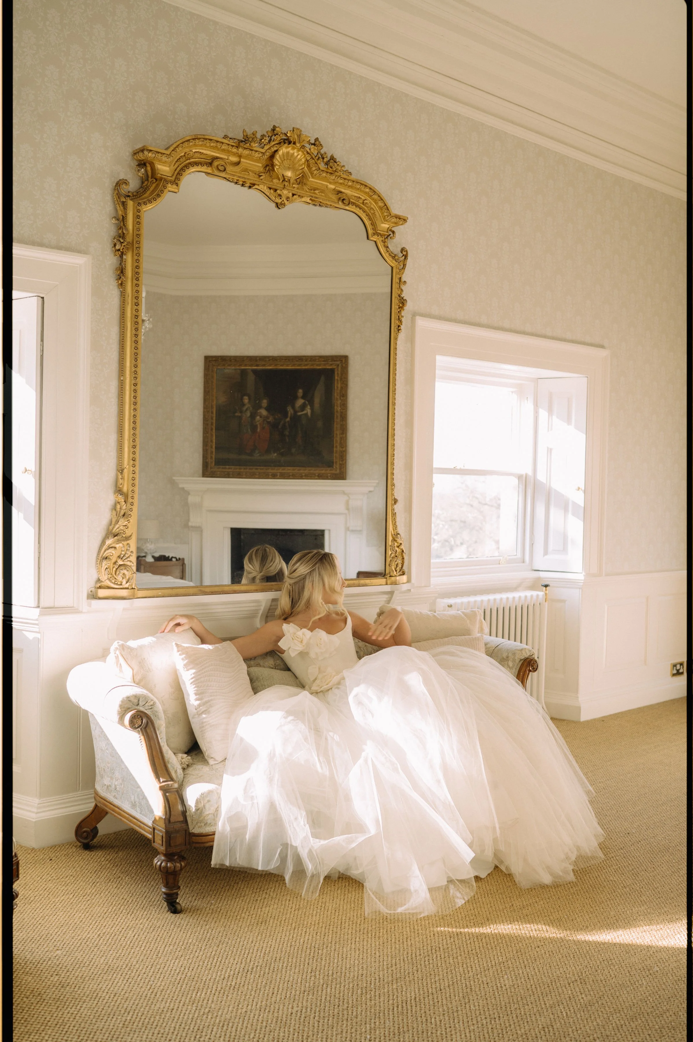 Bride sitting on a vintage sofa in a bright, elegantly decorated room, looking out the window at Earnshill House, Somerset.