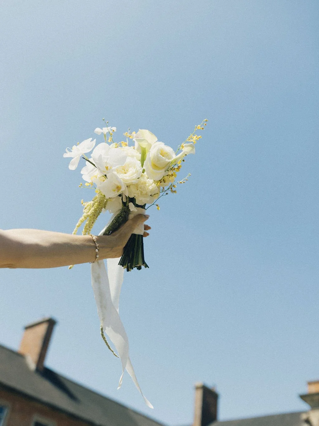 A person holding a bouquet of white flowers against a clear blue sky with rooftops in the background.