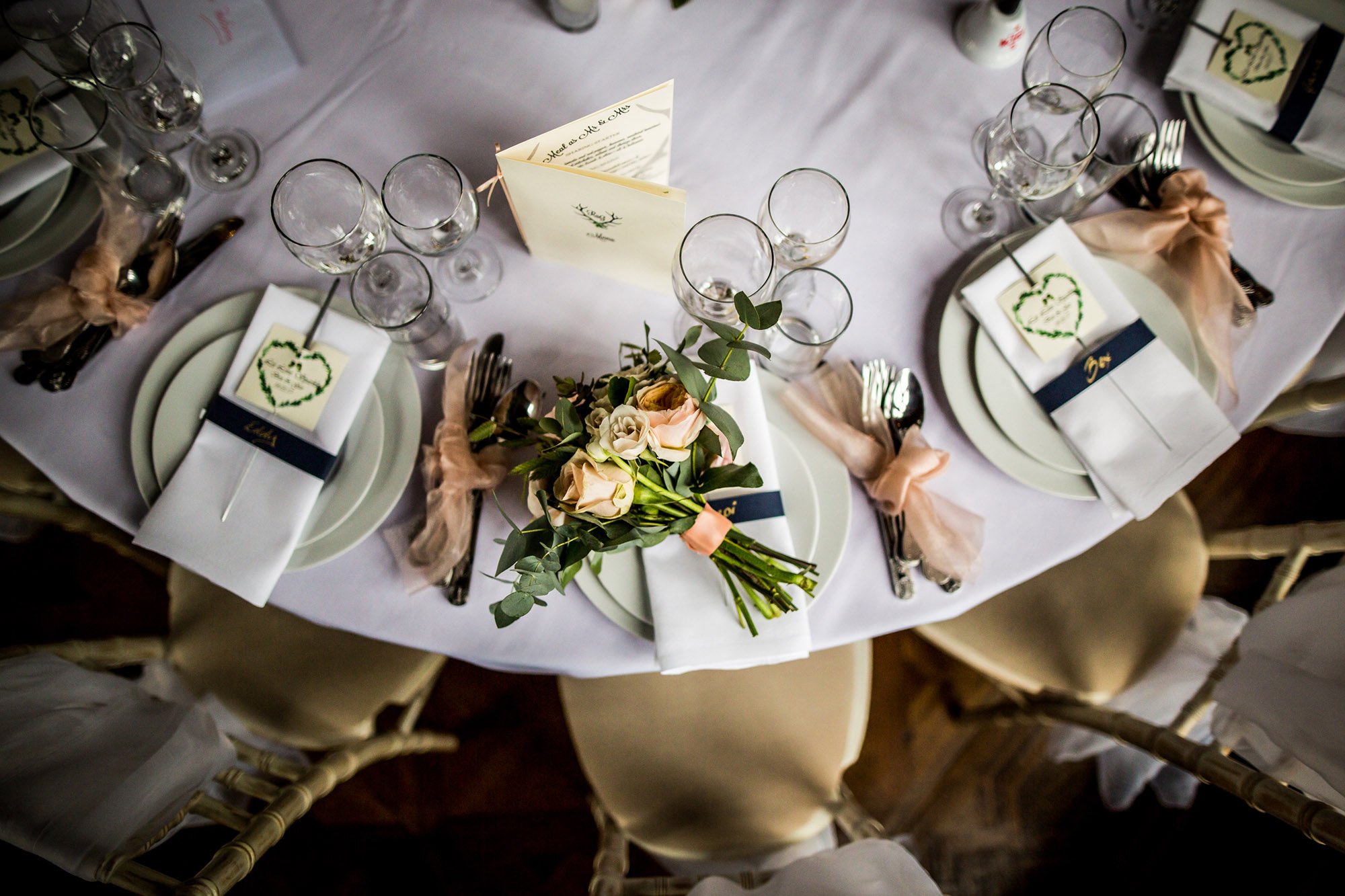 Wedding reception table set with white plates, silverware wrapped with pink ribbons, wine glasses, and a floral centerpiece with roses and greenery. There are printed programs or menus on the table and a folded card with a heart design. The chairs are covered with beige fabric.