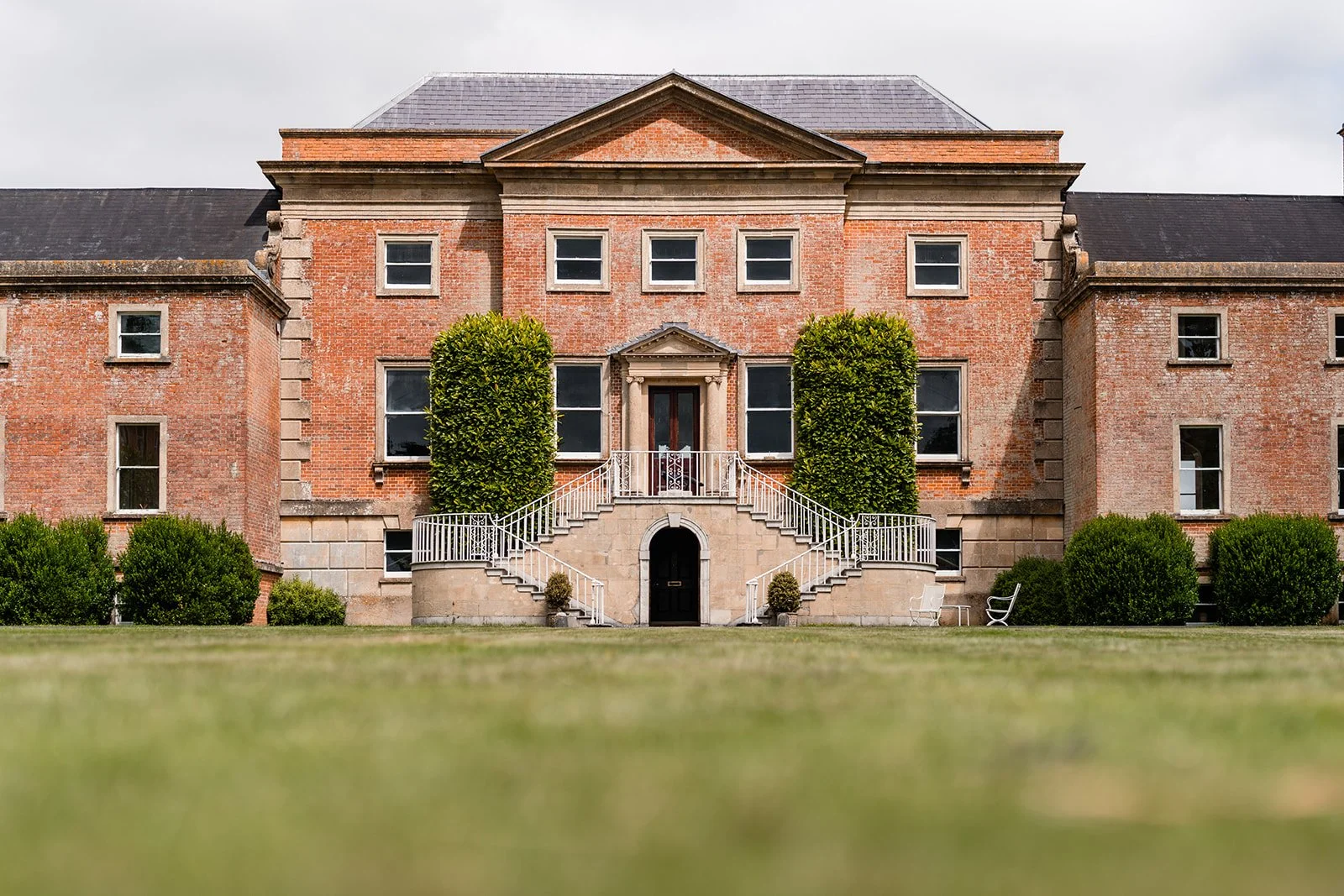 Earnshill House - country mansion with symmetrical landscaping, steps leading up to the entrance, and green bushes and trees surrounding it, under a cloudy sky.