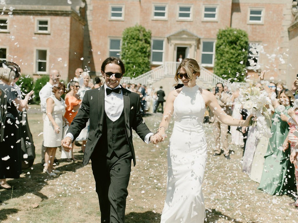 A newly married couple holding hands and smiling while walking through a crowd of guests throwing confetti during their wedding celebration outside Earnshill House in Somerset.