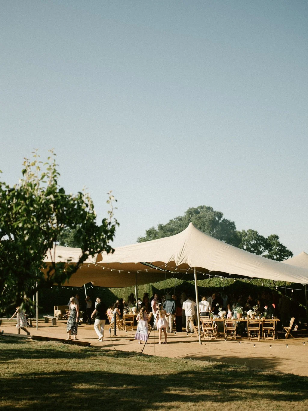 An informal BBQ gathering with people under a large beige tent on a sunny day, with trees and a clear blue sky in the background.
