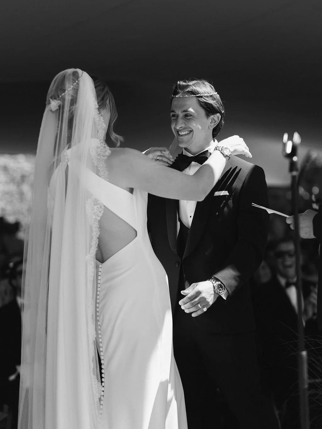 A bride and groom during their wedding ceremony, with the bride adjusting the groom's bow tie, both smiling warmly.