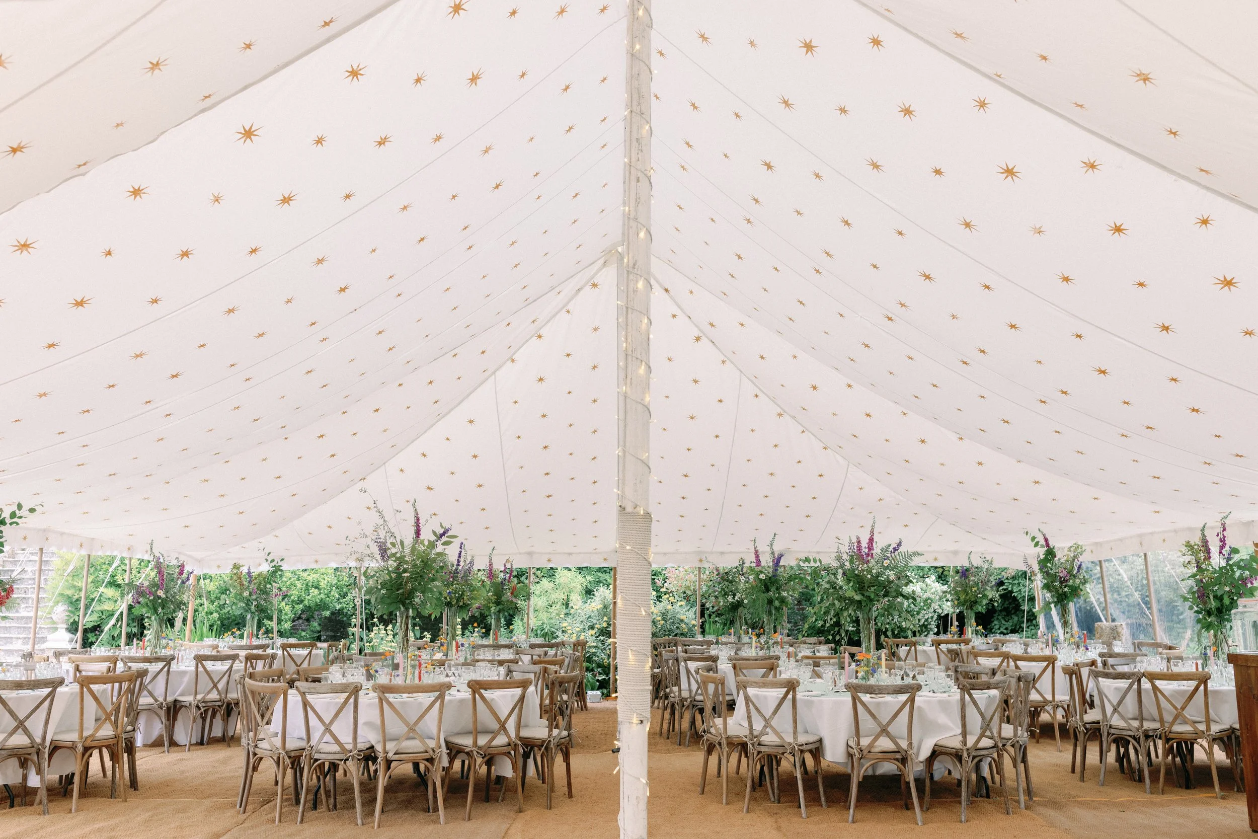 Indoor event space with white dining tents decorated with gold star patterns on the ceiling. There are round tables with white tablecloths, floral arrangements, and wooden chairs. The space is surrounded by greenery and natural light.