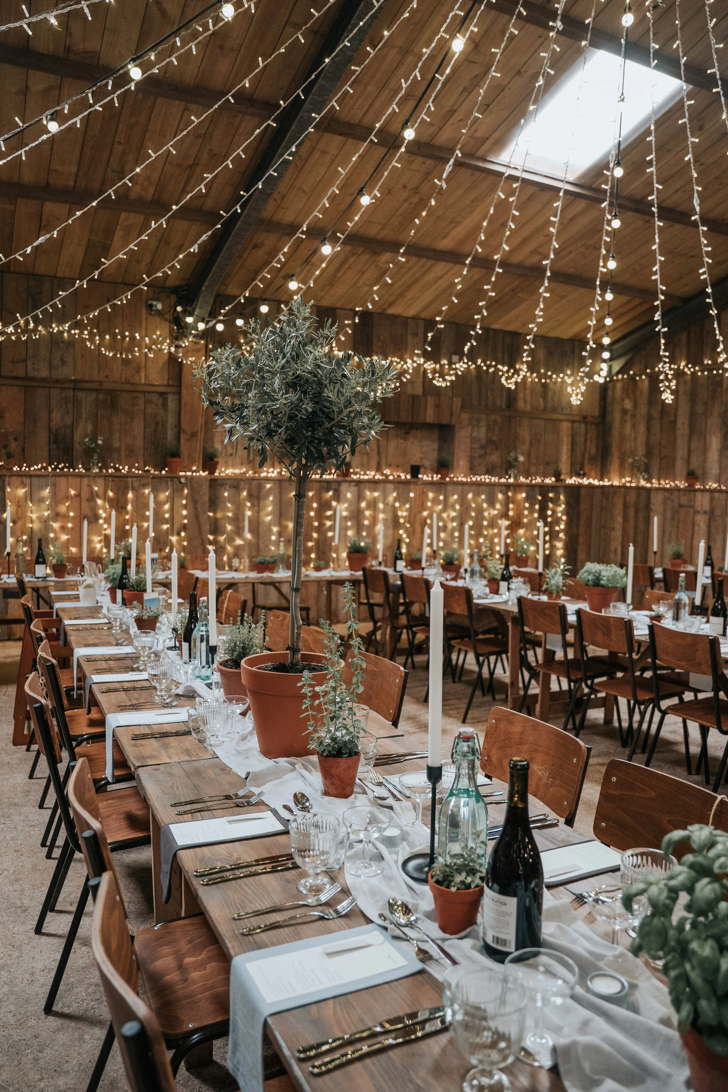 Decorated banquet hall with wooden tables, potted plants, candles, wine bottles, and cutlery, illuminated by string lights and a skylight.