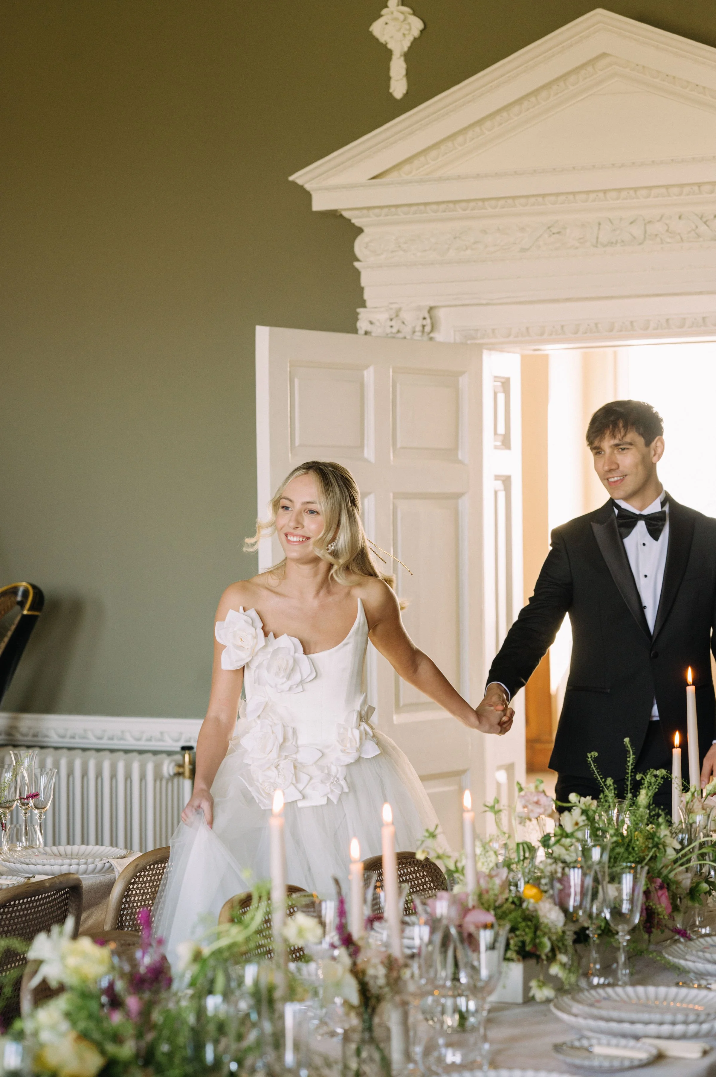 A bride in a white wedding dress with floral details and a groom in a black tuxedo inside a decorated wedding venue, holding hands and smiling.
