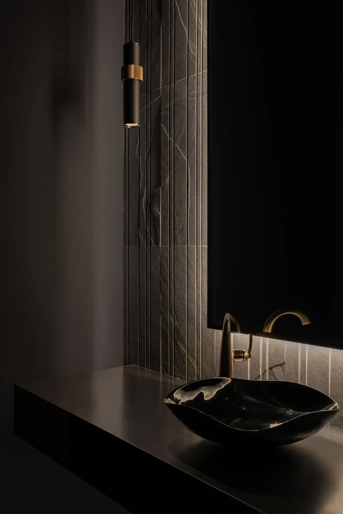 Modern bathroom with a black marble sink, brushed gold faucet, dark stone tiled wall, and a black mirror with backlight, illuminated by a wall-mounted black and gold sconce.