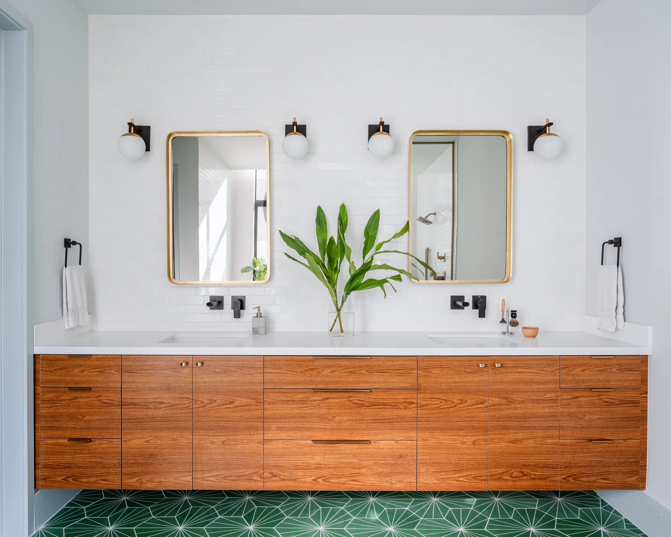 Modern bathroom with double vanity, wooden cabinets, white countertop, two mirrors, black fixtures, green plant, and patterned green floor tiles.