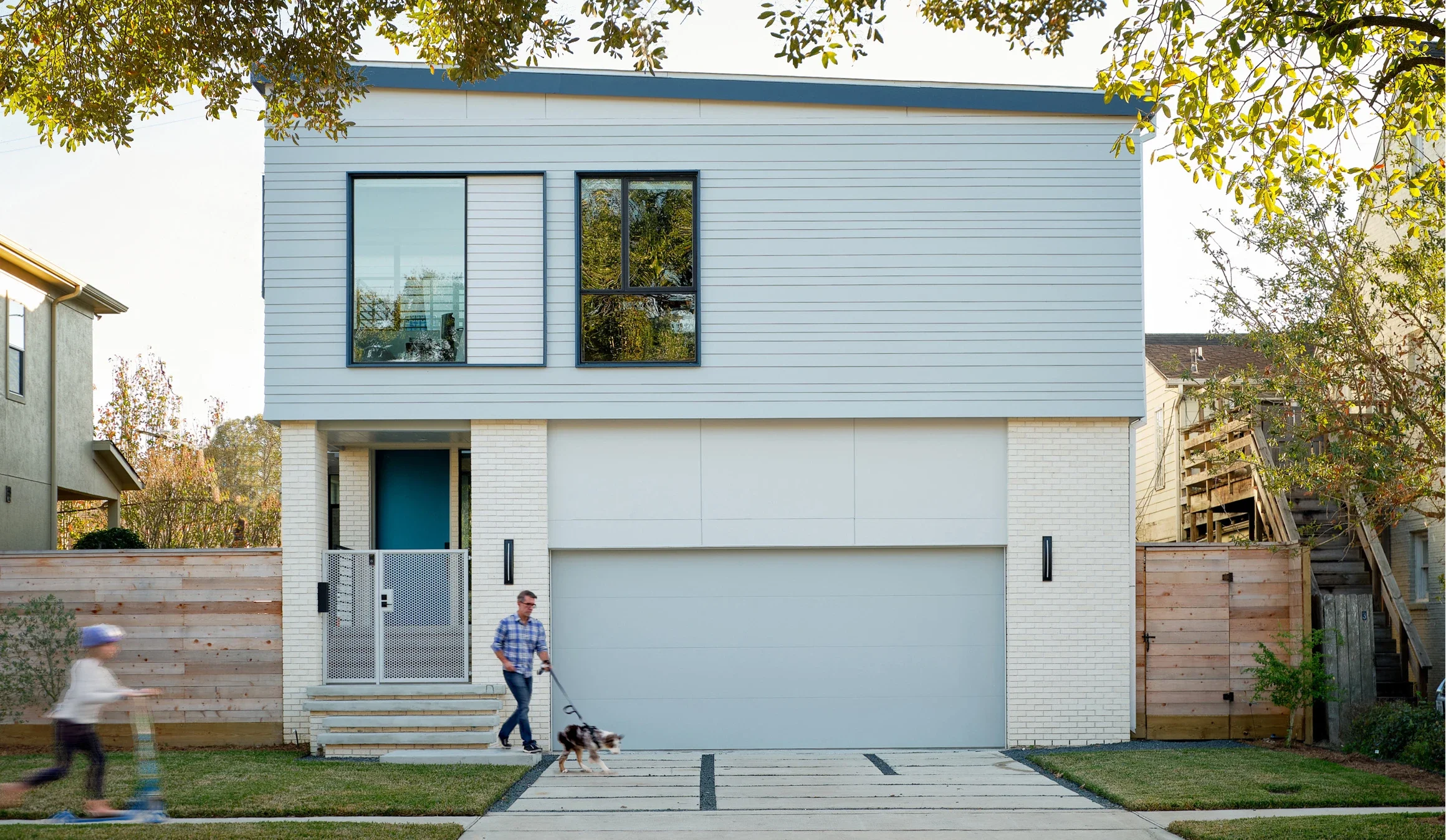 Modern two-story house with white brick and light blue siding, large windows, a driveway, and a wooden fence on both sides. A man walks his dog while a boy rides a scooter in front of the house, with trees and neighboring houses visible.