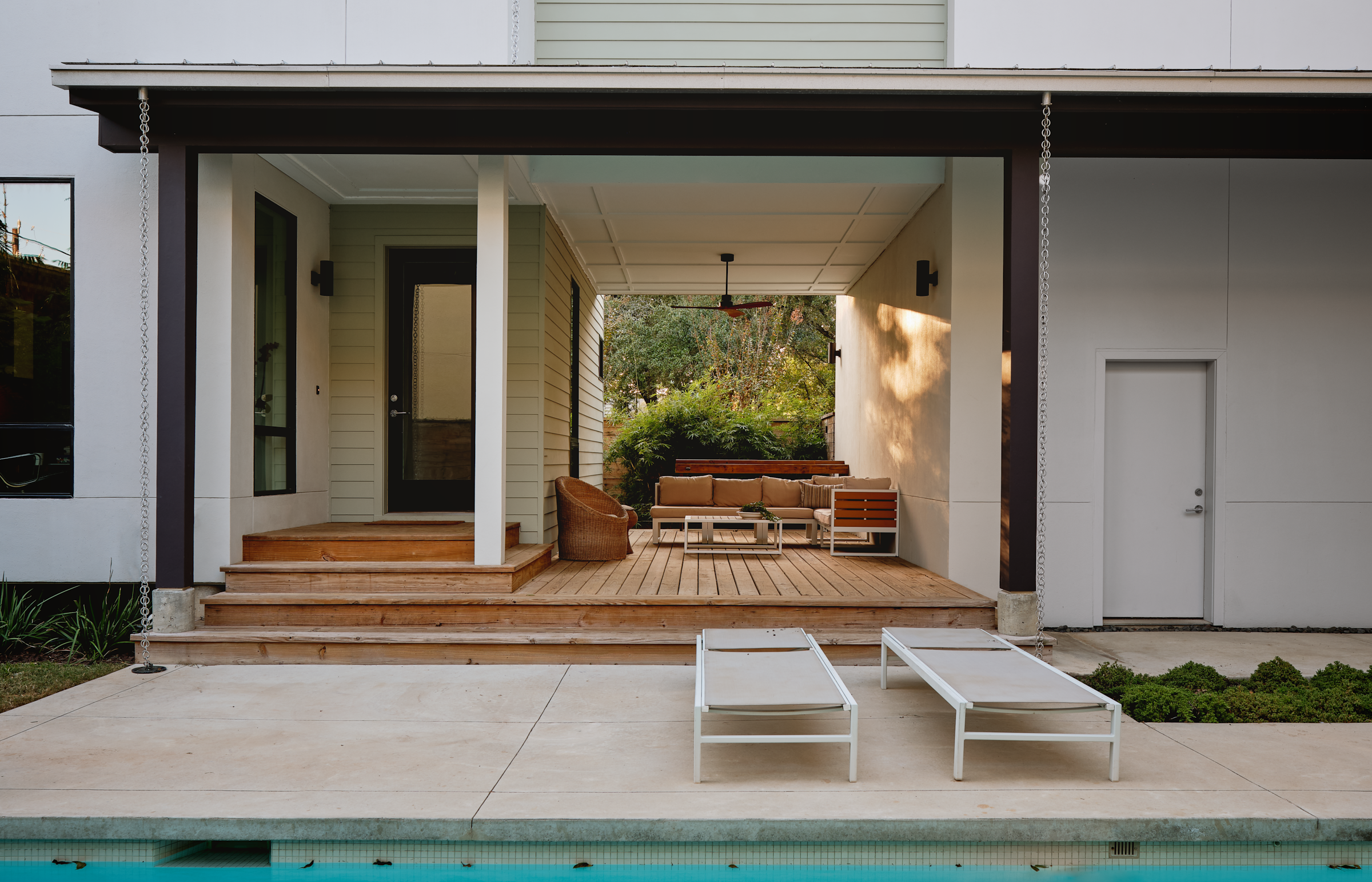 View of a backyard patio with wooden deck, outdoor furniture including a sofa and lounge chairs, surrounded by trees and greenery, with a swimming pool in the foreground.