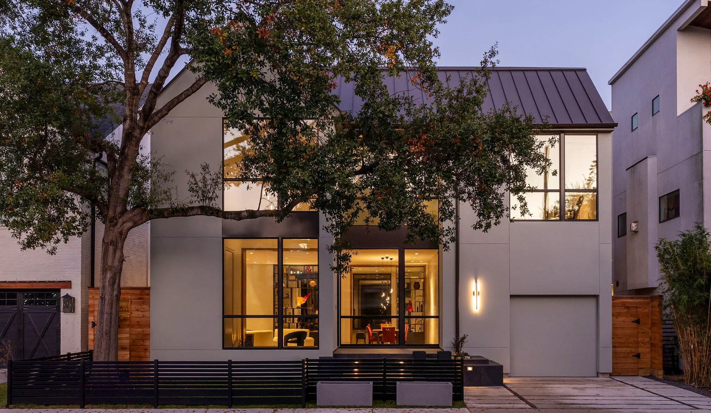 Modern two-story house with large windows, illuminated interior, surrounded by a black fence, with a large tree in the front yard and neighboring homes in an urban setting during dusk.