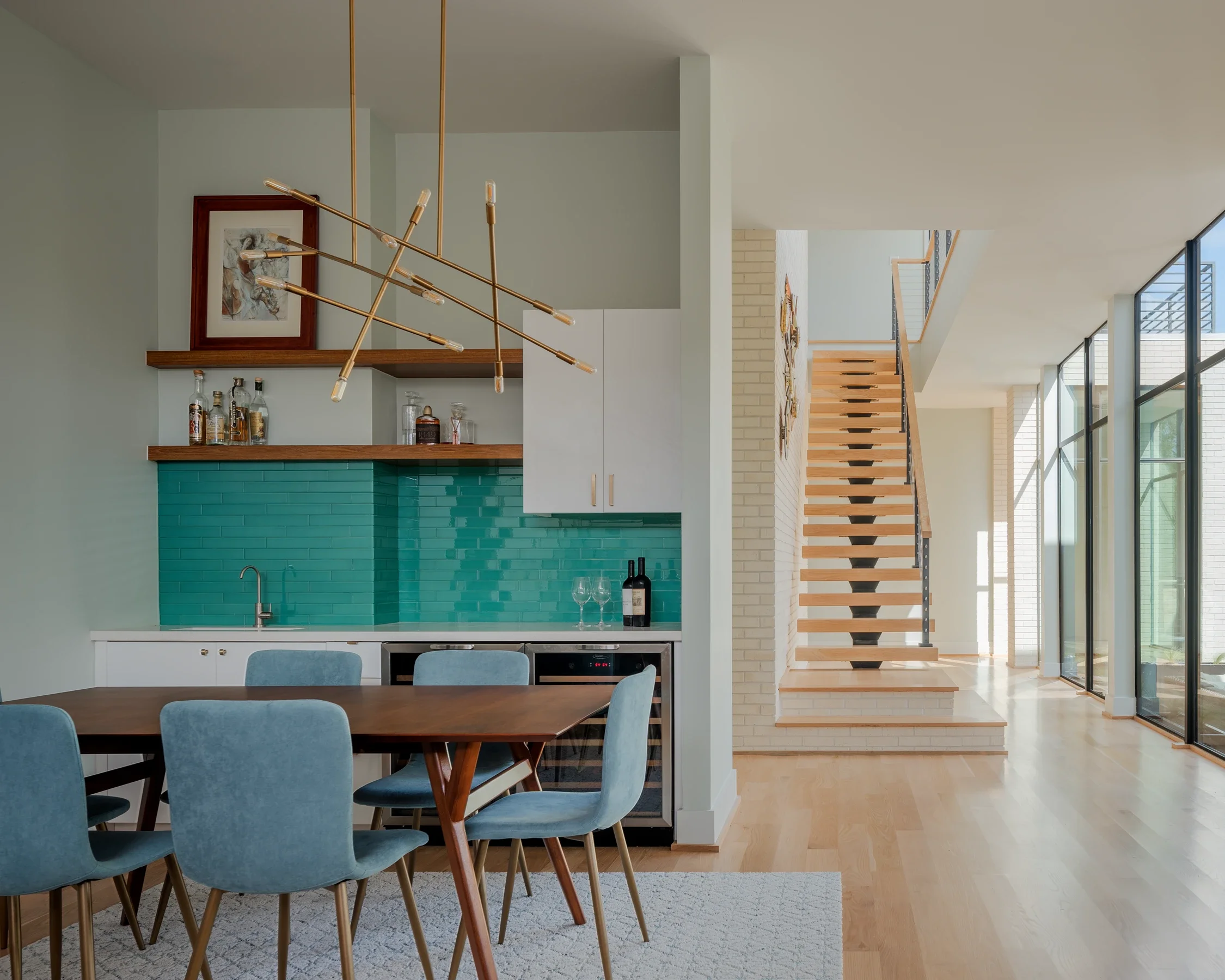 Modern open-concept kitchen and dining area with teal backsplash, wooden staircase, large windows, and contemporary decor.