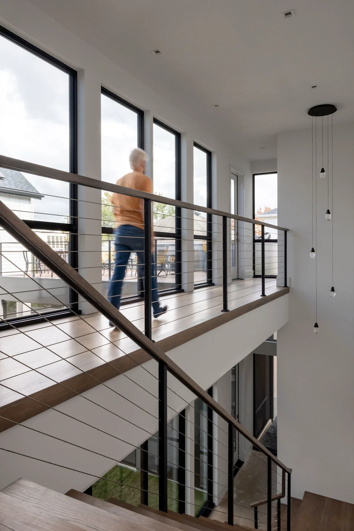 Interior view of a modern house with a staircase and a person walking on the upper level balcony, large floor-to-ceiling windows, white walls, and a minimalist design.