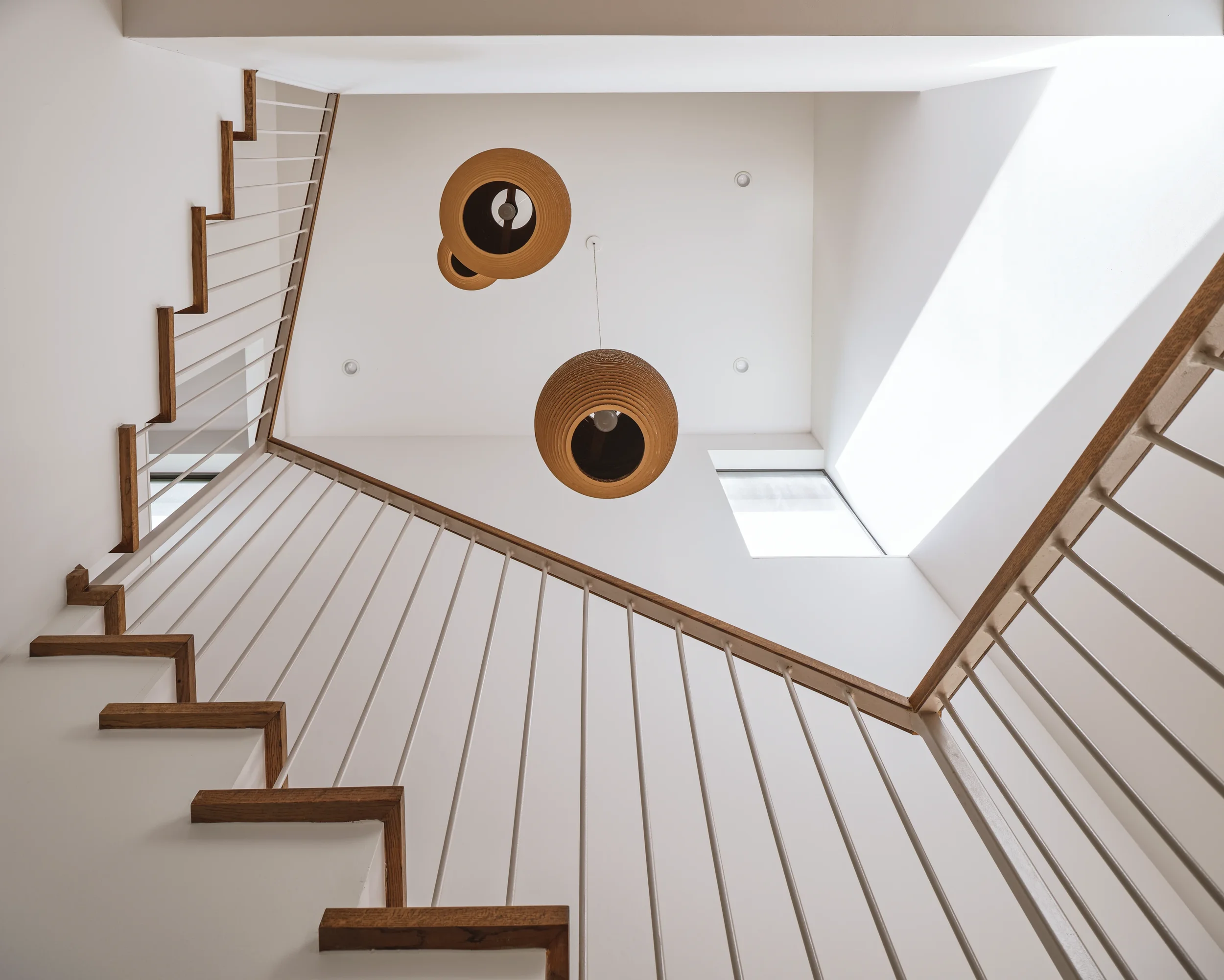 Looking up at a stairwell with white walls, wooden handrails, and two hanging woven pendant lights, with a skylight and a window visible in the ceiling.