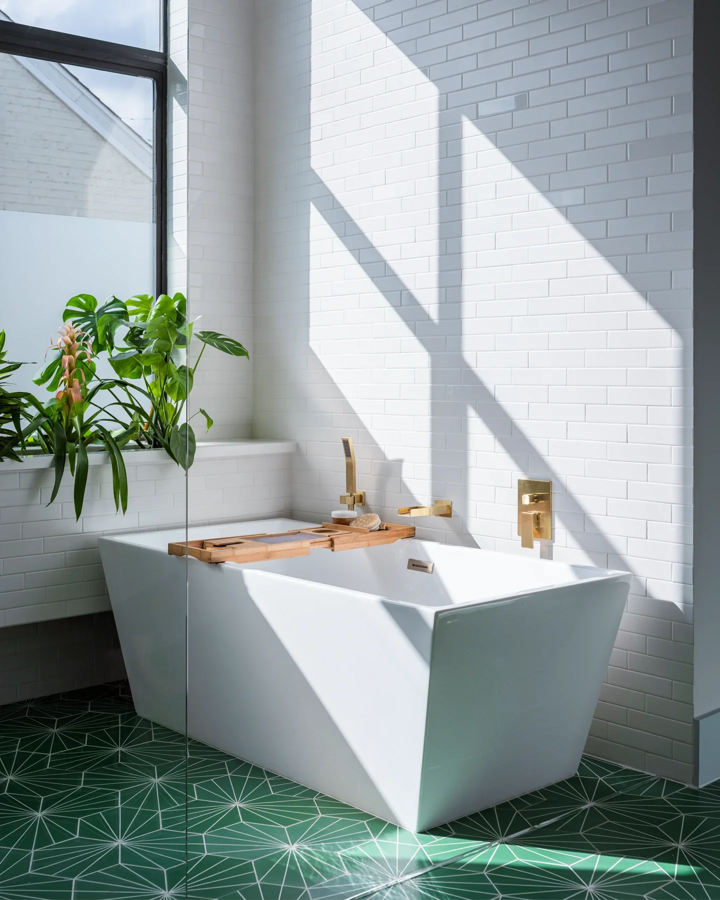 Modern bathroom with white brick walls, a white bathtub, gold fixtures, a wooden bath tray, and a green plant near a large window, with patterned green floor tiles.