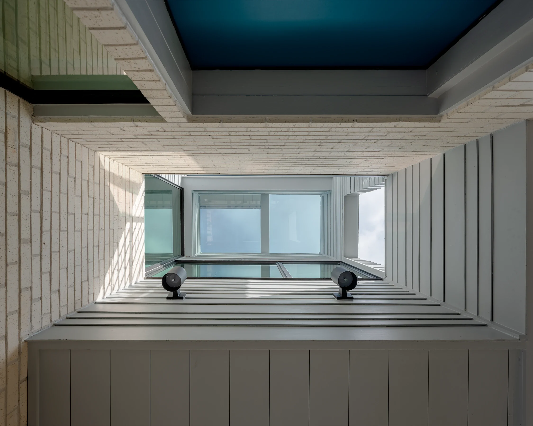 View upward of a modern building's atrium with white brick and metal siding, large windows at the top, and two black security cameras mounted on the wall.