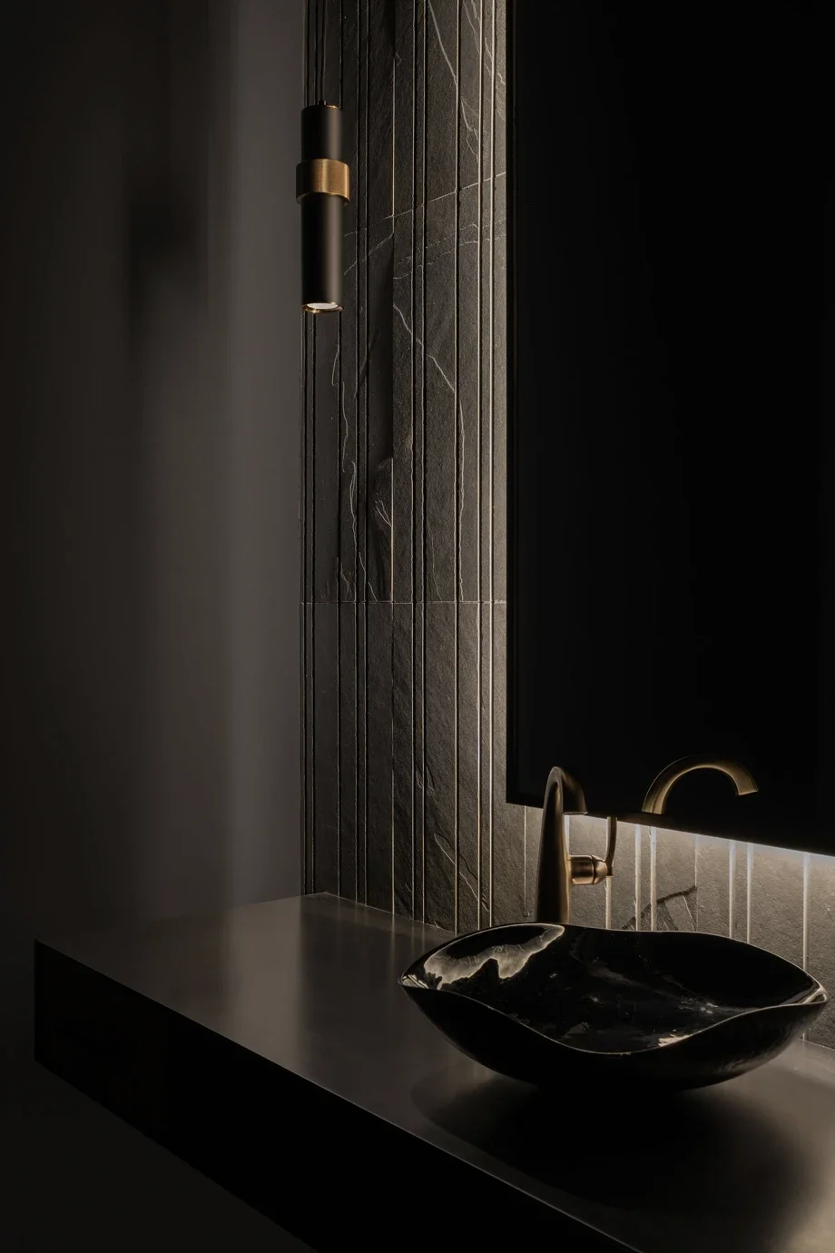 A modern bathroom sink area with a black marble countertop, a black and gold vessel sink, and a gold faucet, with dark textured wall tiles and a large backlit mirror.