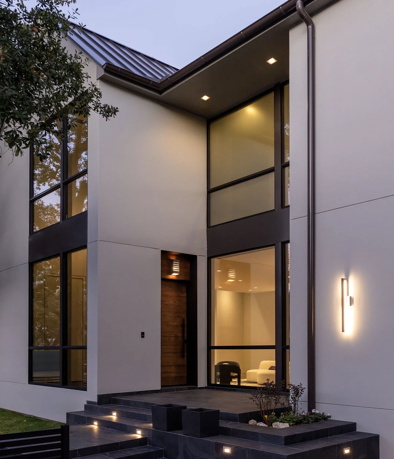 Modern two-story house with large glass windows, a wooden front door, and exterior lighting on a dark tiled entrance with plants.