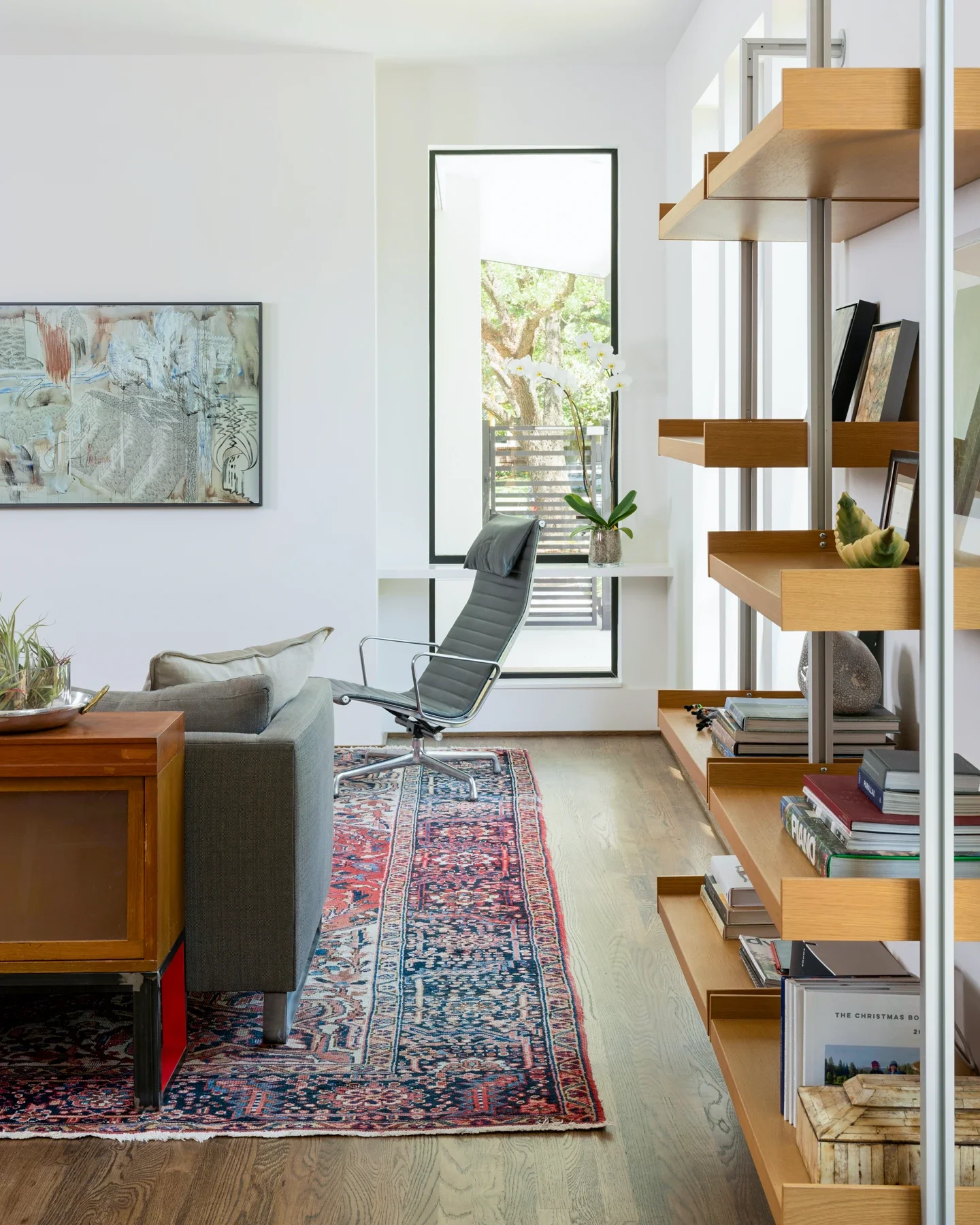 Modern living room with a gray armchair, a wooden side table, a patterned rug, a white bookshelf with books and decorative items, a large window with trees outside, and a piece of abstract art on the wall.