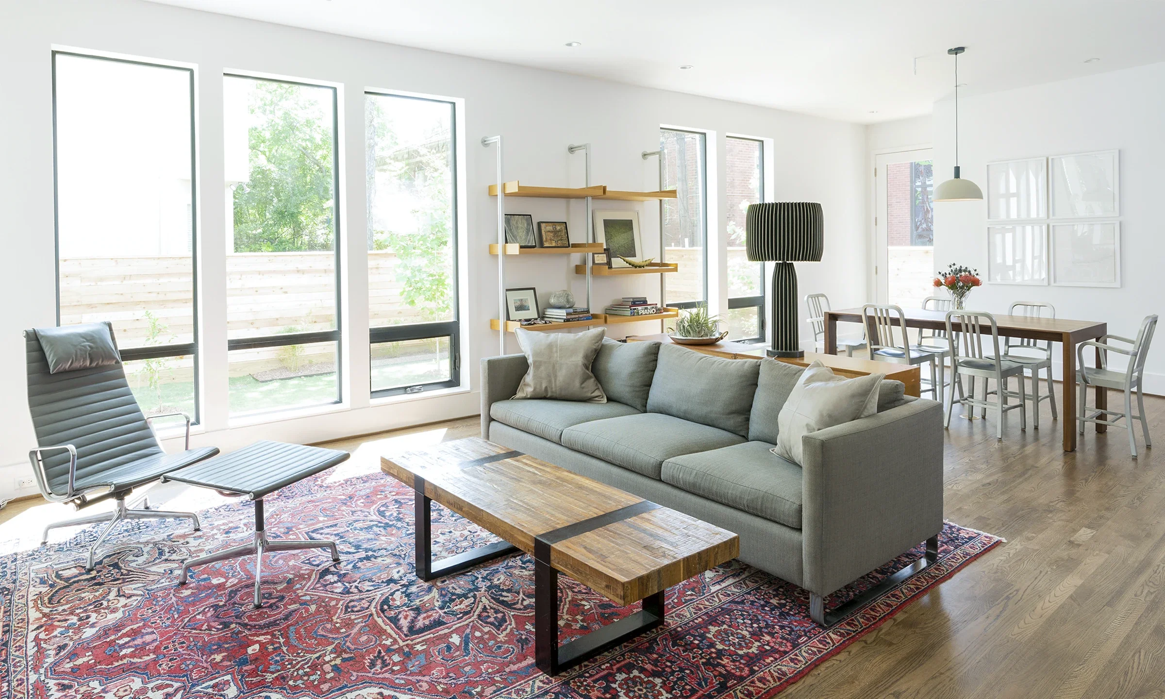 Bright living room with large windows, gray sofa, wooden coffee table, striped armchair, bookshelf, tall black lamp, wooden dining table with six chairs, and wall art.