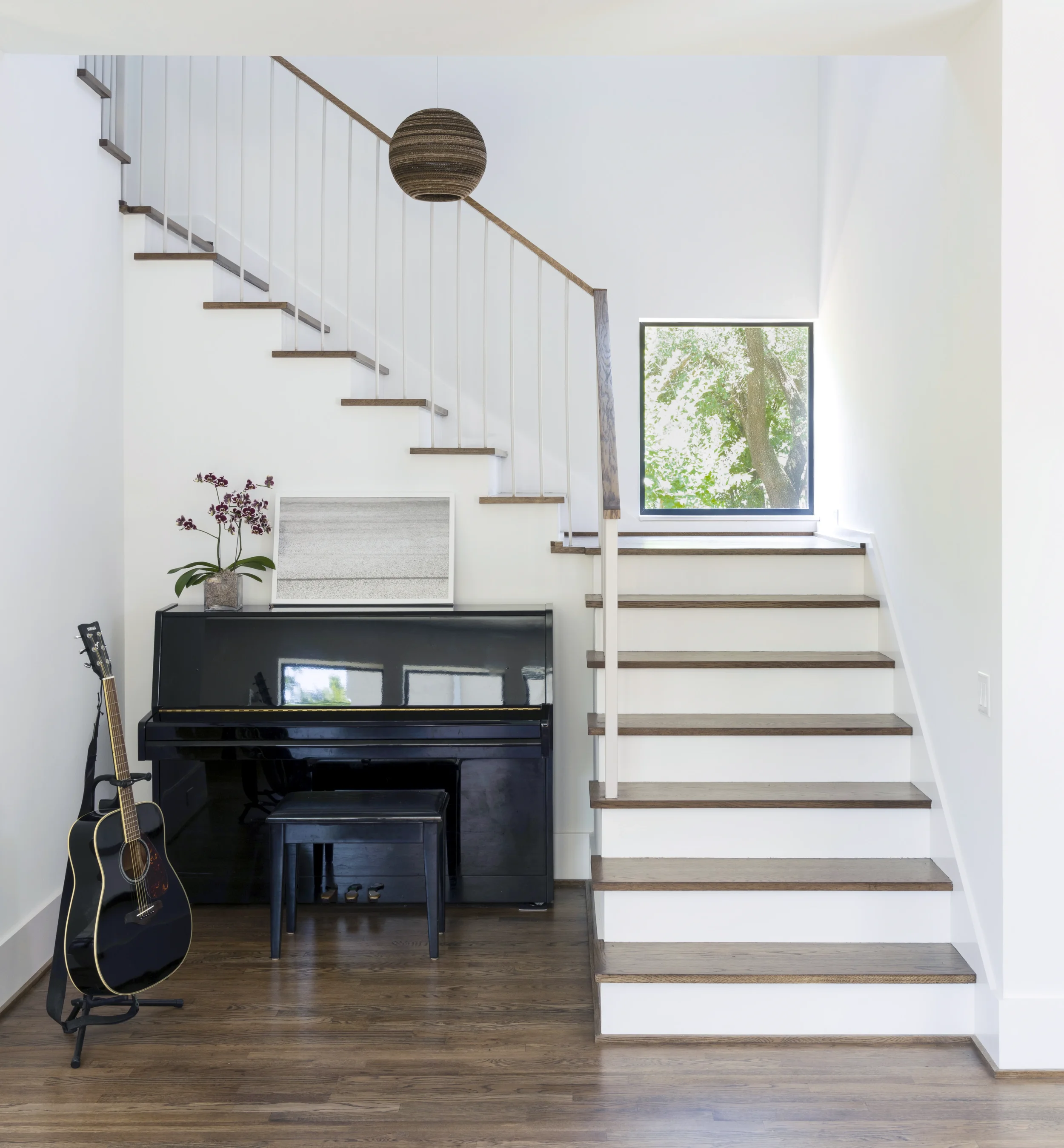 Interior of a modern home with a staircase, a piano, guitar, and a window with greenery outside.