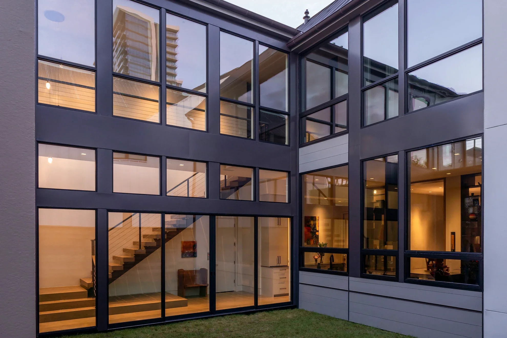 Modern multi-story house with large glass windows and interior lighting visible, including staircase and kitchen area.