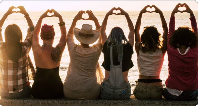 Six women sitting by the water during sunset, making heart shapes with their hands and facing the ocean.