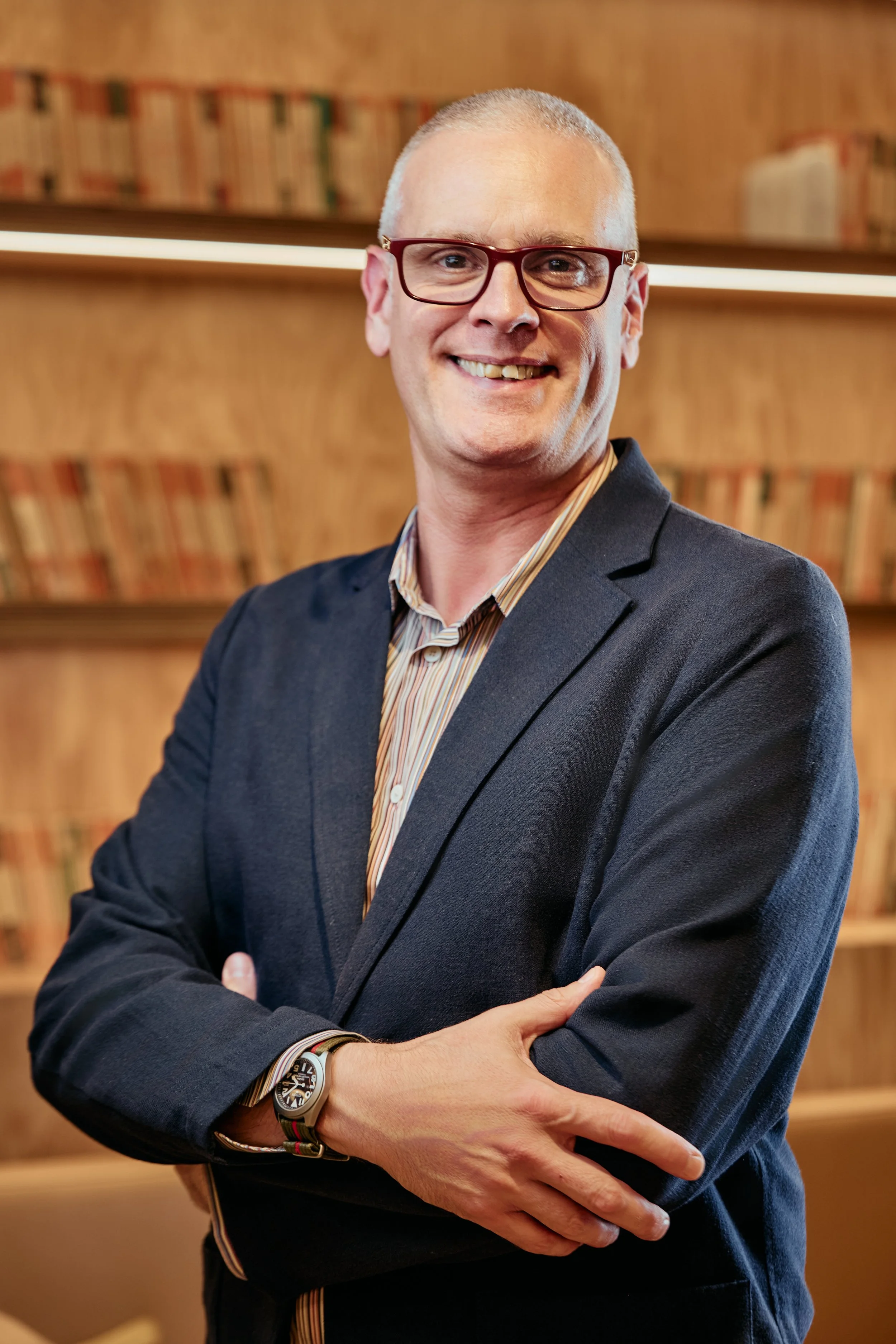 A man with glasses and a closely shaved head, smiling, wearing a dark blazer over a striped shirt, standing in front of a wooden bookshelf filled with books.
