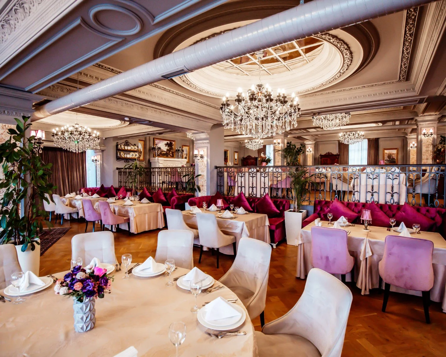 Elegant restaurant interior with chandeliers, pink and cream chairs, tables set with white napkins, glasses, and floral centerpieces, ornate ceiling, and decorative railings.
