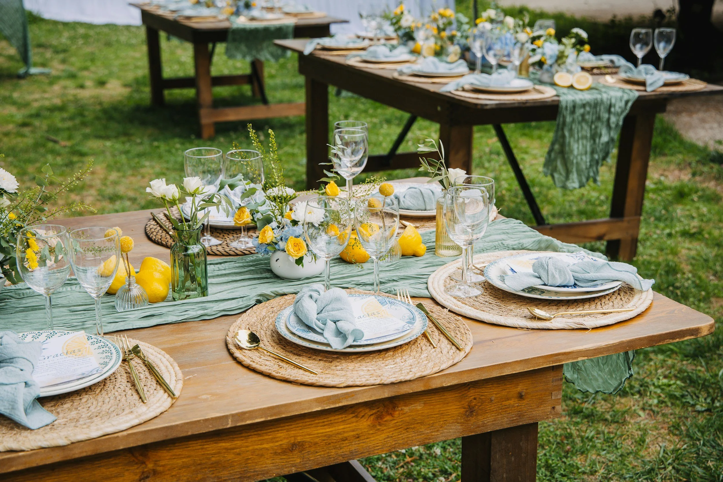 Outdoor dining table set with blue and yellow floral centerpiece, glassware, plates, and napkins on a green table runner, surrounded by wooden tables in a garden setting.