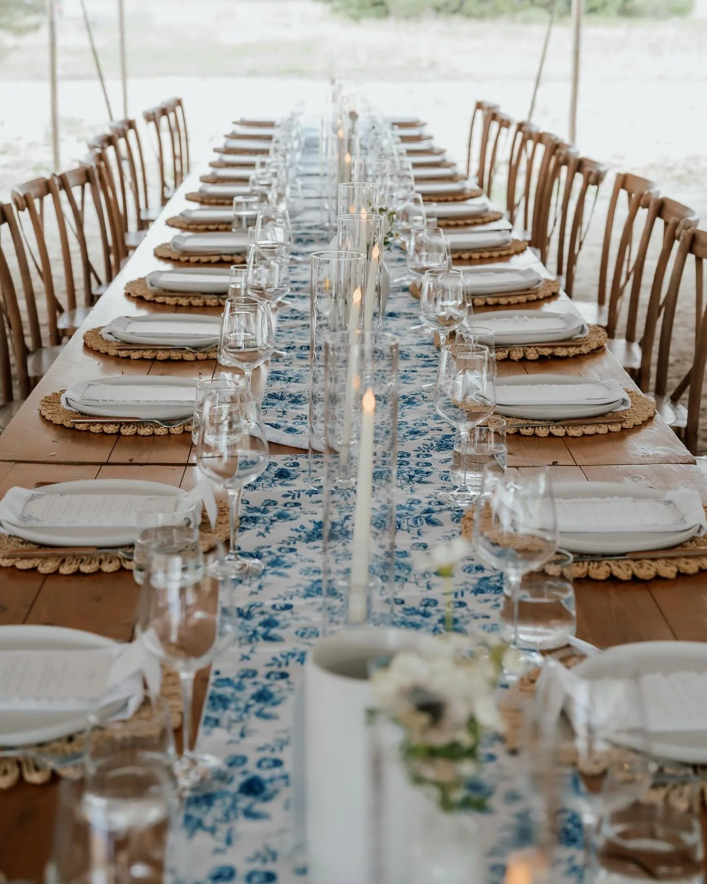 Long dining table set for a formal event with white plates, silverware wrapped in white napkins, clear wine glasses, and tall candles in glass holders, decorated with a blue and white floral table runner and surrounded by wooden chairs.