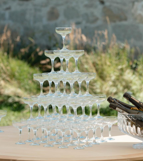 Champagne glasses arranged in a pyramid with a glass of champagne at the top, outdoors with greenery and a stone wall in the background.