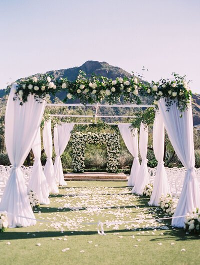 Outdoor wedding altar with white drapes and floral arrangements, set against a mountainous backdrop.