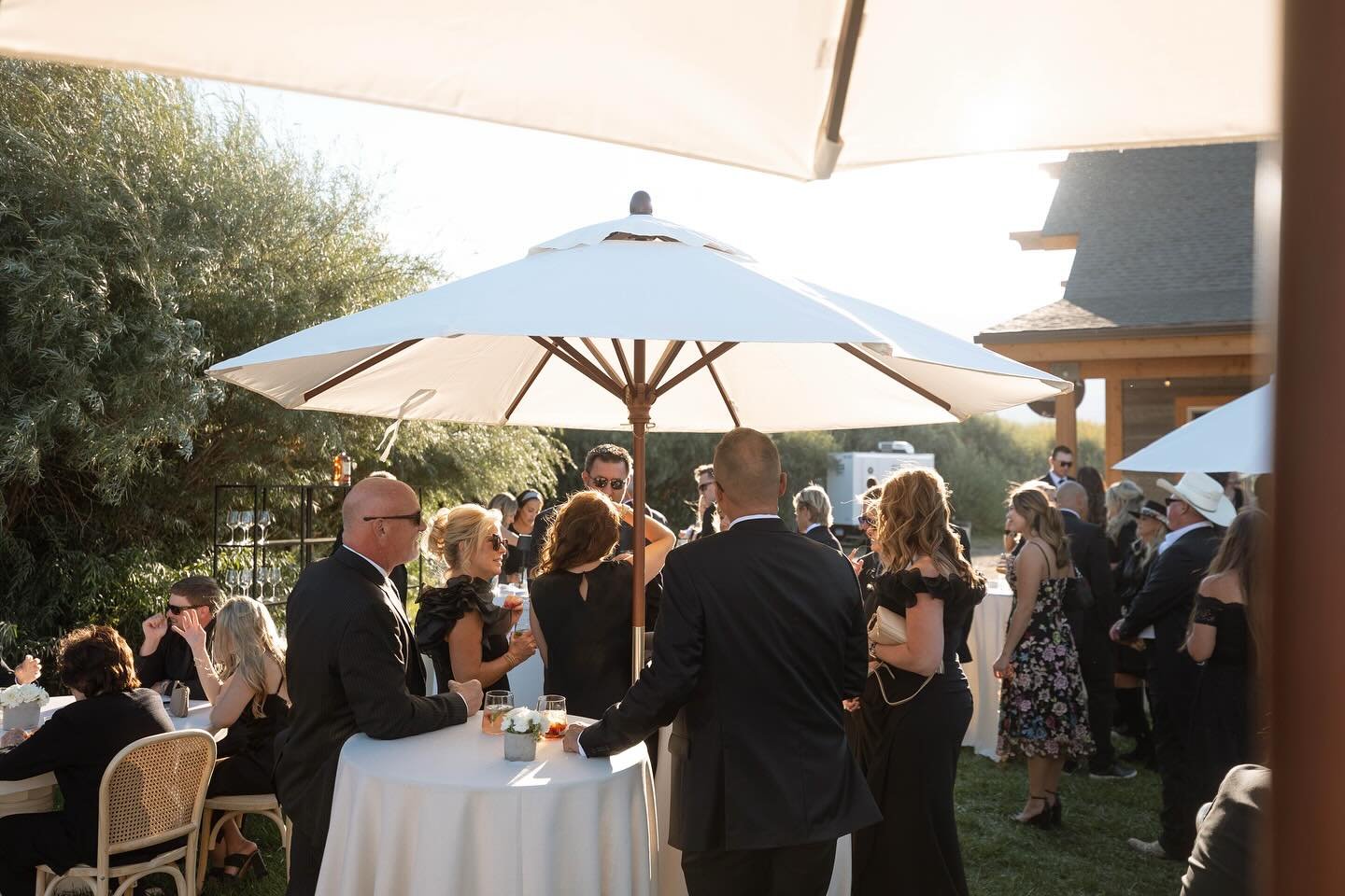 People in formal attire gathered outdoors at a social event, standing around tables with white tablecloths and large umbrellas for shade, in a garden-like setting.