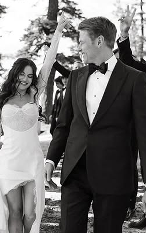 A wedding celebration outdoors with a bride and groom holding hands, smiling, and surrounded by guests making celebratory gestures, in a wooded area.