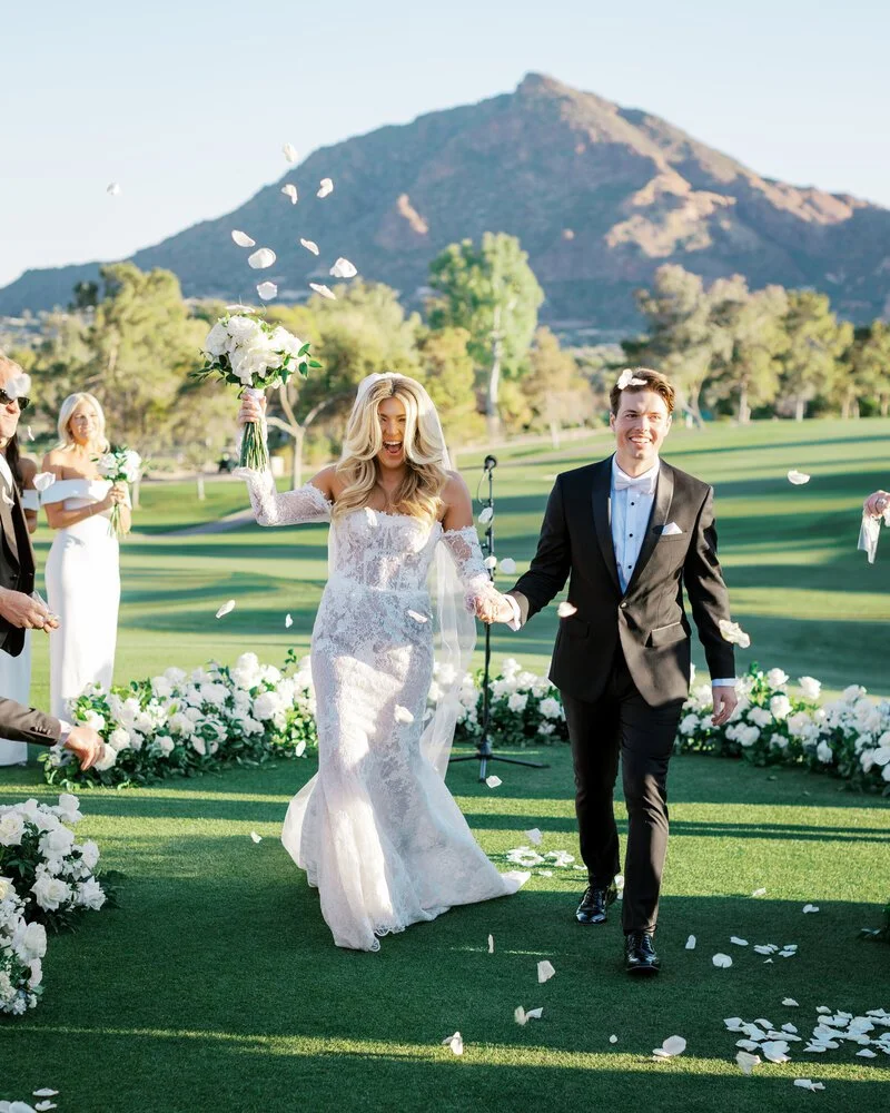 A bride and groom walking hand in hand down an outdoor wedding aisle, surrounded by white flowers, with a mountain in the background, during daylight. The bride is holding a bouquet and the groom is smiling.