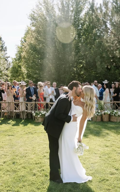A newlywed couple sharing a kiss at their outdoor wedding ceremony, with guests watching in the background on a sunny day.