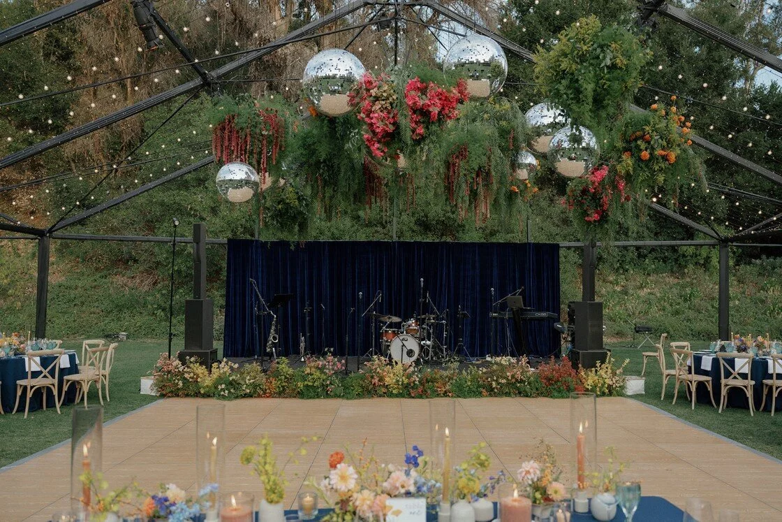 Outdoor event space with a stage set for a musical performance, decorated with hanging greenery and large reflective disco balls, surrounded by tables with floral arrangements and candles.