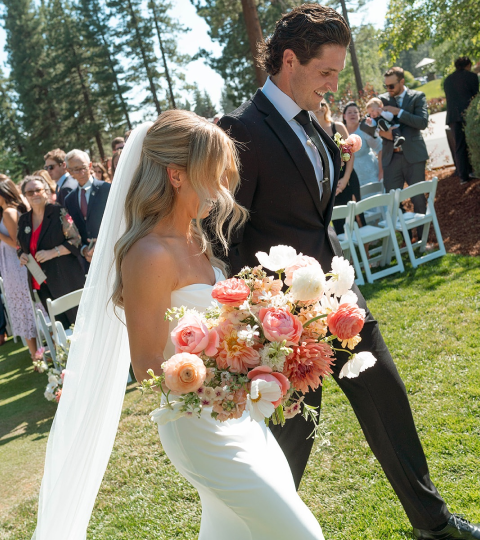 Bride and groom walking outdoors at wedding, surrounded by seated guests and trees