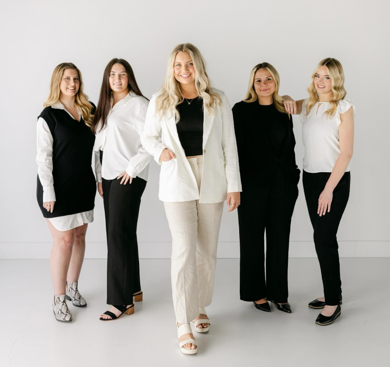 Five women standing in a row against a plain white background, posing and smiling. The central woman is wearing a white blazer and cream-colored pants, while the others are dressed in black and white outfits.