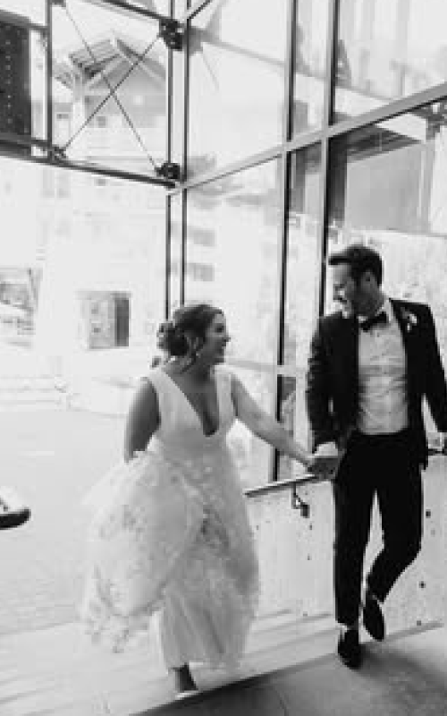 Bride and groom holding hands and smiling inside a modern glass building.