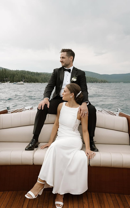A bride and groom sitting on a boat, with the groom sitting on the back of the seat and the bride sitting in front of him, against a backdrop of water and forested mountains under a cloudy sky.