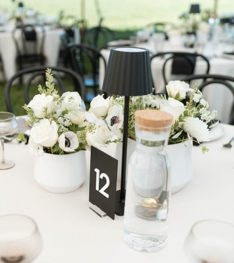Table centerpiece with white flowers, black table number sign showing 12, black lamp, and water pitcher on a dining table in a restaurant setting