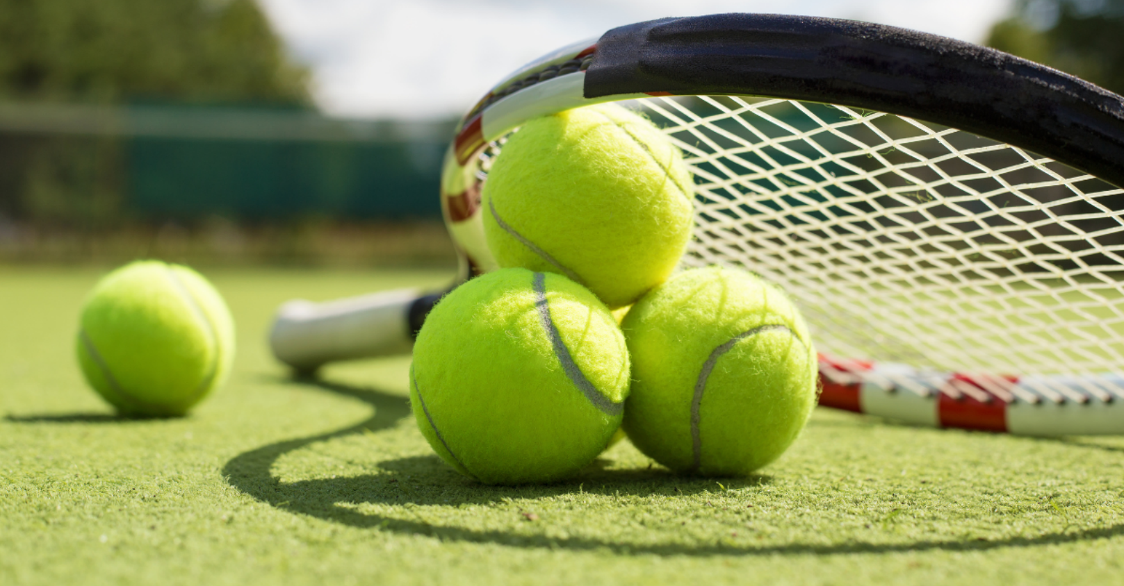 Four bright yellow tennis balls on a tennis court near a tennis racket and net, with a lush green background.